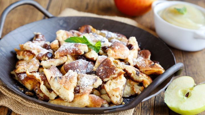 Auf einem dunklen Teller liegt fluffiger Kaiserschmarrn, best&auml;ubt mit Puderzucker, umgeben von einem gr&uuml;nen Apfel und Apfelmus.