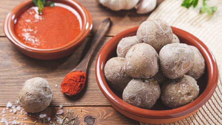 Kleine, runde Knollen liegen in einer Sch&uuml;ssel neben einer w&uuml;rzigen Salsa und frischen Zutaten auf einem Holztisch.