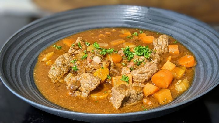 Eine Sch&uuml;ssel Eintopf mit Rindfleisch, Karotten und Petersilie, serviert in einem modernen, dunklen Teller auf einem Tisch.