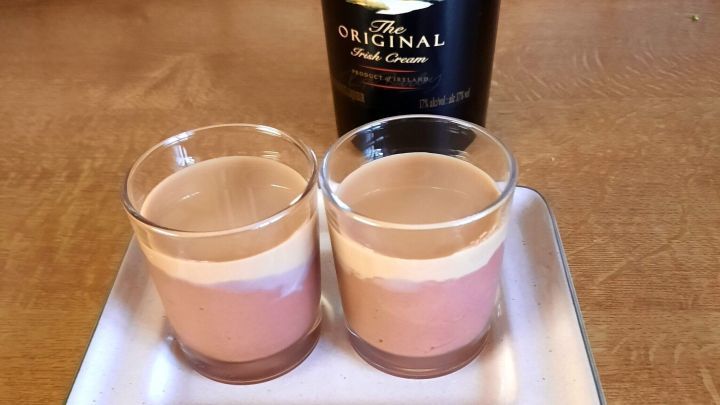 Zwei Gl&auml;ser Schokopudding-Dessert mit Baileys stehen auf einem wei&szlig;en Teller, umgeben von einer Holzoberfl&auml;che.