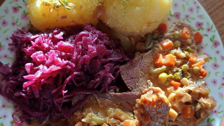 Sauerbraten mit Gem&uuml;se und Rotkohl liegt auf einem blumengemusterten Teller, umgeben von zwei gro&szlig;en Kartoffeln.