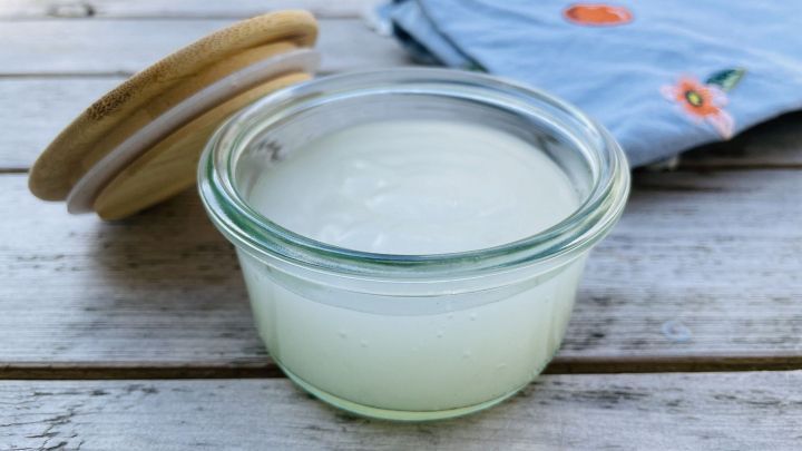 Ein Glas mit selbstgemachtem Kleister steht auf einem Holztisch, w&auml;hrend ein gesticktes Tuch im Hintergrund sichtbar ist.