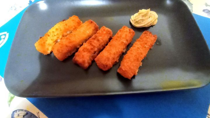 Auf einem schwarzen Teller liegen goldbraun gebratene Fischst&auml;bchen, neben einem Klecks Sauce in einem bunten Umfeld.