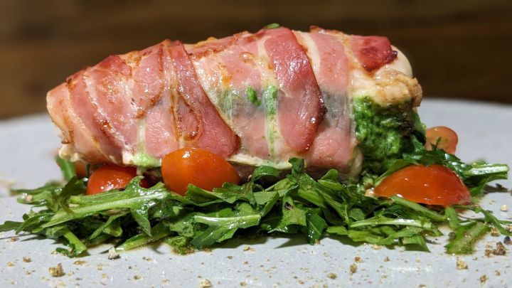 Gef&uuml;llte H&auml;hnchenbrust umwickelt mit Speck liegt auf frischem Rucola und Cherrytomaten, angerichtet auf einem grauen Teller.