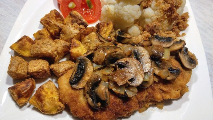 R&ouml;stkartoffeln liegen knusprig auf einem Teller, kombiniert mit paniertem Fleisch, Pilzen, Blumenkohl und Tomatenscheiben.