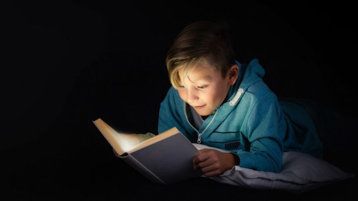 Ein Junge liegt auf einem Kissen und liest gespannt ein Buch, umgeben von Dunkelheit, die die Leselampe betont.
