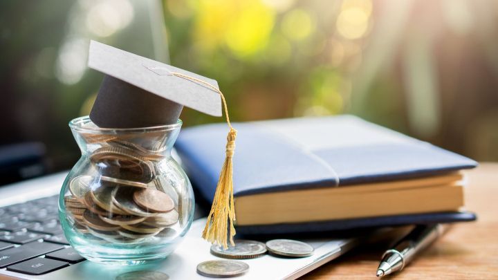 Ein Glas voller M&uuml;nzen mit einem kleinen Absolventenhut steht auf einem Laptop neben einem ge&ouml;ffneten Buch im Sonnenlicht.