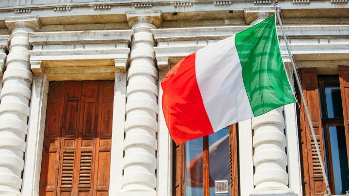 Eine italienische Flagge weht vor einem historischen Geb&auml;ude mit wei&szlig;en S&auml;ulen und h&ouml;lzernen Fenstern an einem sonnigen Tag.