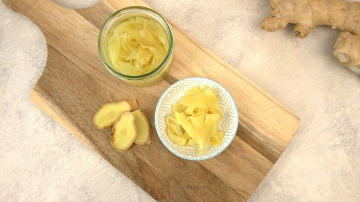 Eingelegter Ingwer liegt in einer Schale auf einem Holzbrett neben einem Glas, umgeben von frischem Ingwer.