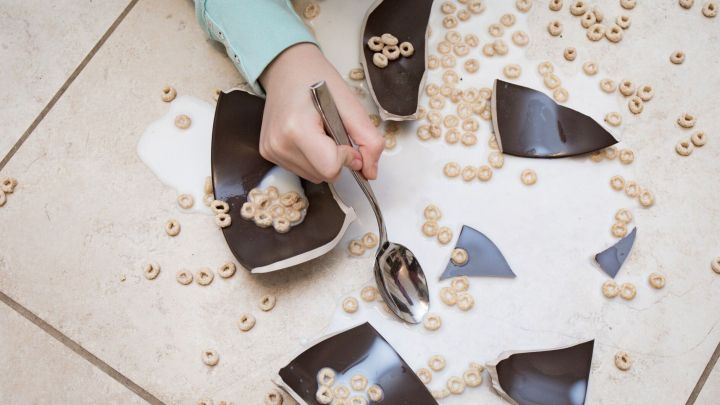 Ein Arm greift nach einem L&ouml;ffel, w&auml;hrend zerbrochene Sch&uuml;sselst&uuml;cke und Cerealien auf dem Boden verteilt sind.