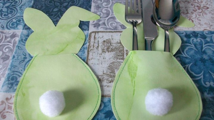 Zwei gr&uuml;ne Bestecktaschen in Hasenoptik halten Besteck und sind dekorativ auf einem bunten Tischmuster platziert.