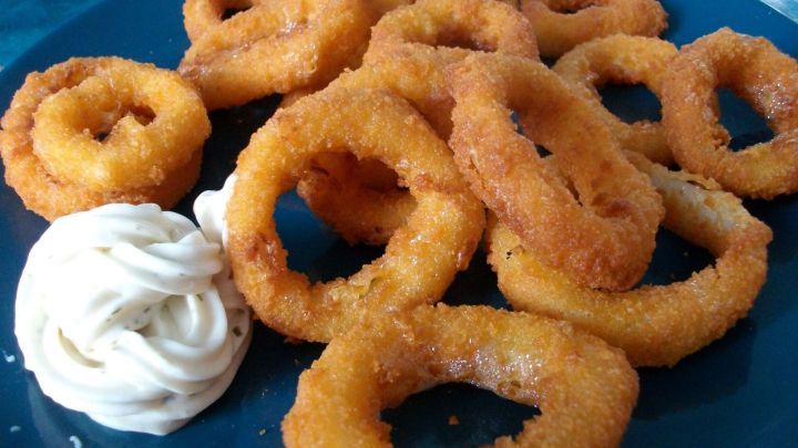 Krosse Calamari-Ringe liegen auf einem blauen Teller neben einer Portion Mayonnaise in einem hellen Umfeld.