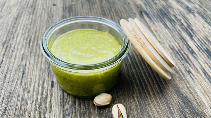 Eine gr&uuml;ne Pistaziencreme steht in einem Glasbeh&auml;lter auf einer Holzoberfl&auml;che, umgeben von Pistazienschalen.
