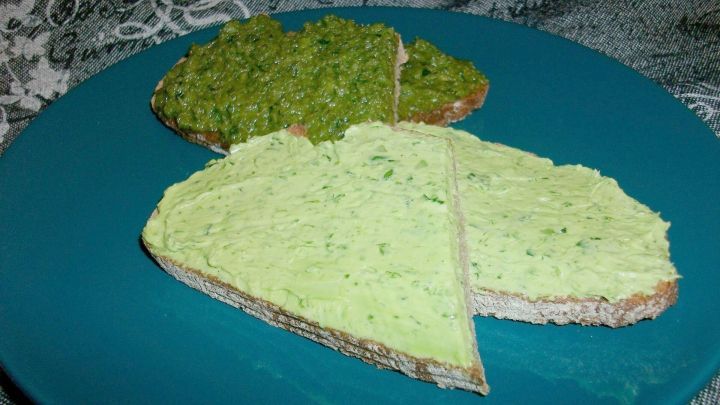 Zwei belegte Brote mit B&auml;rlauchbutter und einer gr&uuml;nen Paste liegen auf einem blauen Teller in einem rustikalen Umfeld.