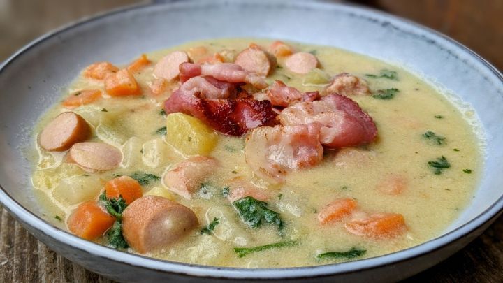 Ein cremiger Gem&uuml;seeintopf mit Wurst und Speck liegt in einer Sch&uuml;ssel, umgeben von einem rustikalen Holztisch.