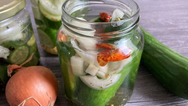 Eingemachte Gurken in einem Glas mit Zwiebel und getrockneten Tomaten, dazu frische Gurke und Zwiebel auf dem Tisch.