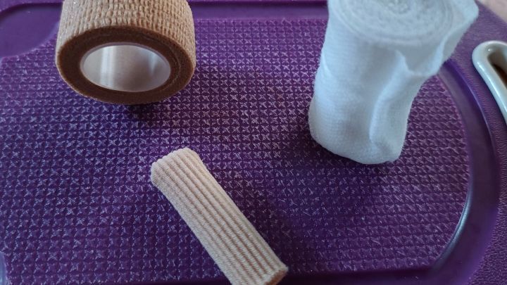 Ein Verbandmaterial liegt auf einer lila Unterlage, w&auml;hrend ein Pflaster und ein Watteverband nebeneinander platziert sind.