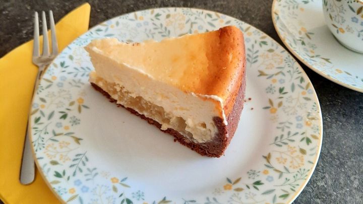 Ein St&uuml;ck K&auml;se-Apfelmuskuchen auf einem blumigen Teller, neben einer Gabel und einer Tasse auf einer dunklen Tischoberfl&auml;che.