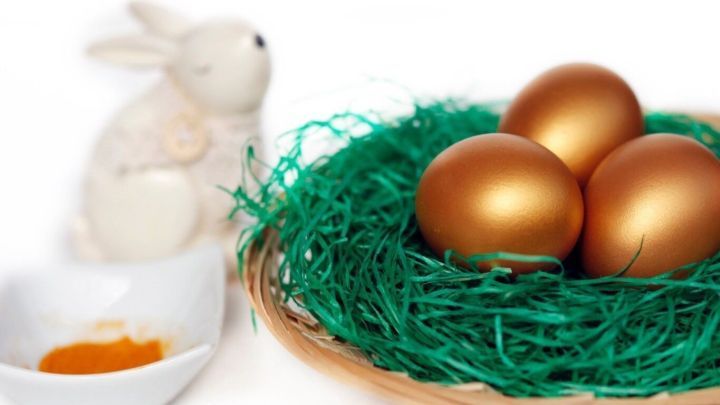 Goldene Ostereier liegen in einem Nest aus gr&uuml;nem Gras, w&auml;hrend ein Hase daneben steht und eine kleine Sch&uuml;ssel mit Farbe zeigt.