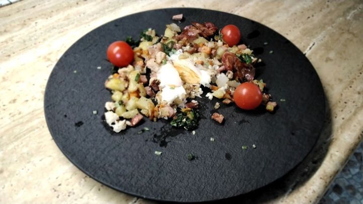 Auf dem schwarzen Teller liegt eine Gem&uuml;sepfanne mit Ei, die frisch angerichtet ist und mit Kirschtomaten garniert wurde.