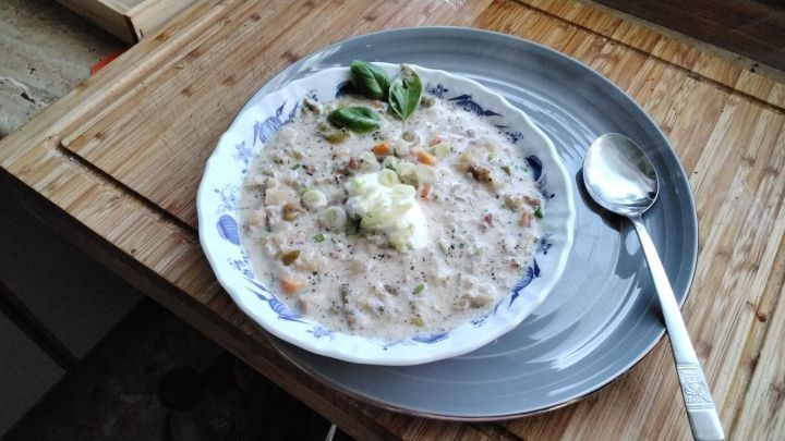 Cremige Gem&uuml;sesuppe wird in einer tiefen Sch&uuml;ssel serviert, umgeben von frischen Kr&auml;utern auf einem Holztisch.