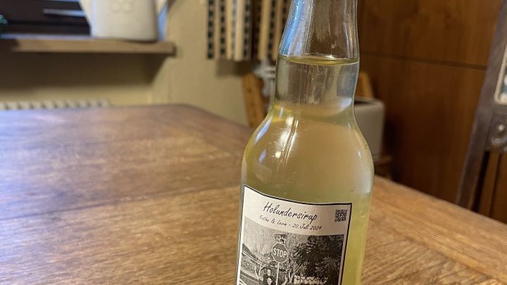 Eine Flasche Holunderbl&uuml;tensirup steht auf einem Holztisch, daneben ein Holzfahrrad, umgeben von einer gem&uuml;tlichen Wohnatmosph&auml;re.