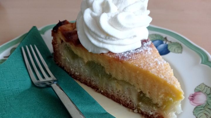 Ein St&uuml;ck Obstkuchen mit saurer Sahne und Sahneh&auml;ubchen liegt auf einem Teller neben einer Gabel auf einem Holztisch.