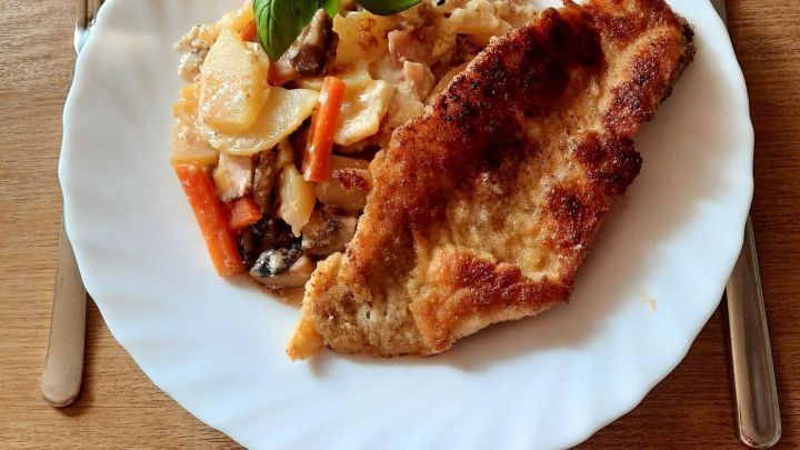 Paniertes Rotbarschfilet liegt neben einer bunten Gem&uuml;sebeilage auf einem wei&szlig;en Teller, dekoriert mit Kirschtomaten und Basilikum.