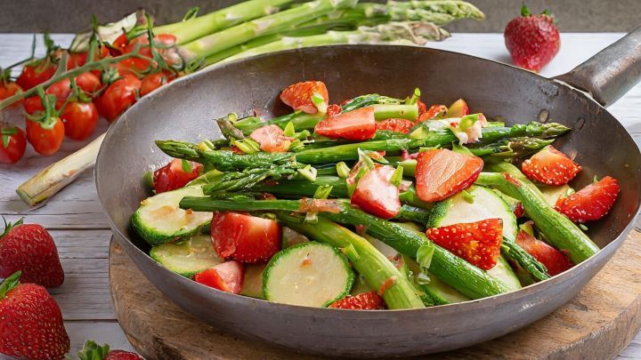 Eine bunte Gem&uuml;sepfanne mit gr&uuml;nem Spargel und Erdbeeren liegt in einer Pfanne, umgeben von frischem Gem&uuml;se und Fr&uuml;chten.