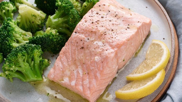 Zartes Lachsfilet liegt neben dampfgegarten Brokkoli und Zitronenscheiben auf einem Teller, umgeben von neutraler Tischdecke.