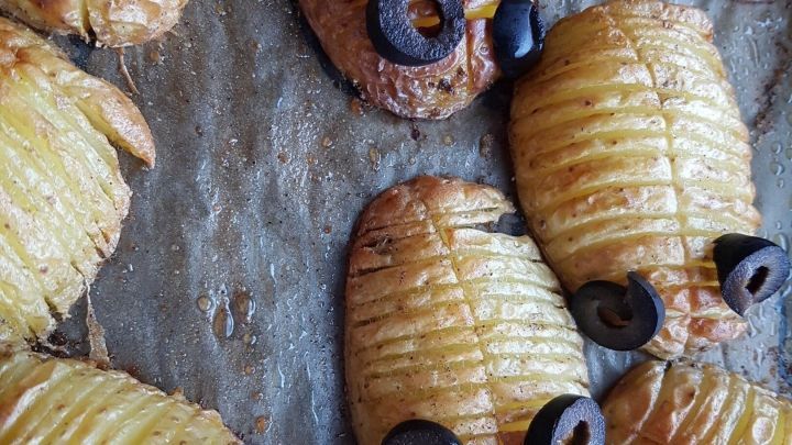Hasselback-Kartoffeln liegen gebacken auf einem Blech, dekoriert mit Oliven, die wie K&auml;fer-Augen aussehen.