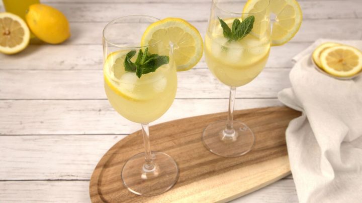Zwei Gl&auml;ser Limoncello Spritz mit Minze und Zitronenscheiben stehen auf einem Holzbrett, umgeben von Zitronen.