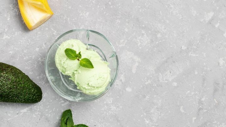 Zwei L&ouml;ffel cremiges Bananen-Avocado-Eis liegen in einer Sch&uuml;ssel, umgeben von einer Avocado und einem St&uuml;ck Banane auf grauem Untergrund.