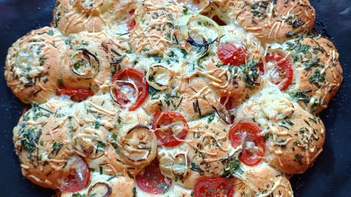 Eine runde, gebackene Pizzablume mit K&auml;se und frischem Gem&uuml;se liegt auf einem schwarzen Teller und sieht appetitlich aus.