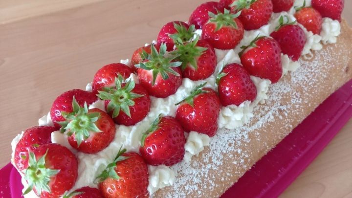 Eine Erdbeer-Biskuitrolle liegt auf einem pinken Teller, dekoriert mit frischen Erdbeeren und Sahne.