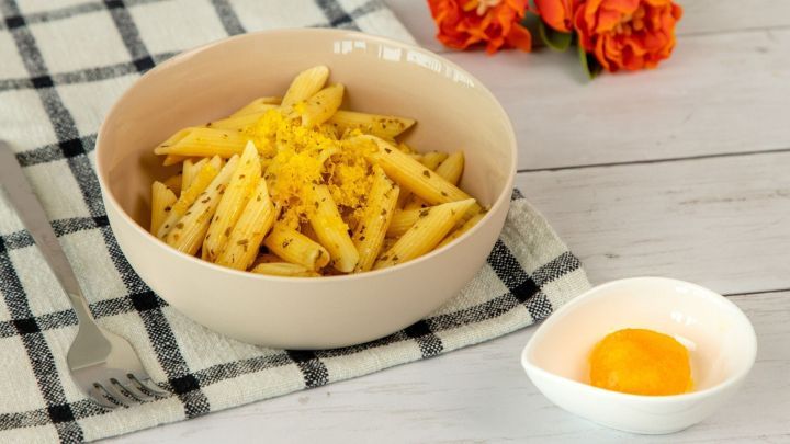 Pasta liegt in einer Sch&uuml;ssel, garniert mit gebeiztem Eigelb, daneben ein gelbes Eigelb in einer kleinen Schale, Blumen im Hintergrund.