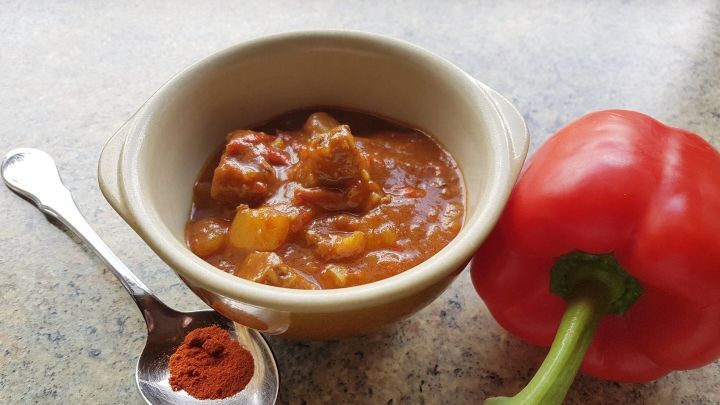 Ein Sch&uuml;ssel Gulasch mit St&uuml;ckchen im Innenbereich, daneben ein roter Paprika und eine L&ouml;ffel mit Paprikapulver.