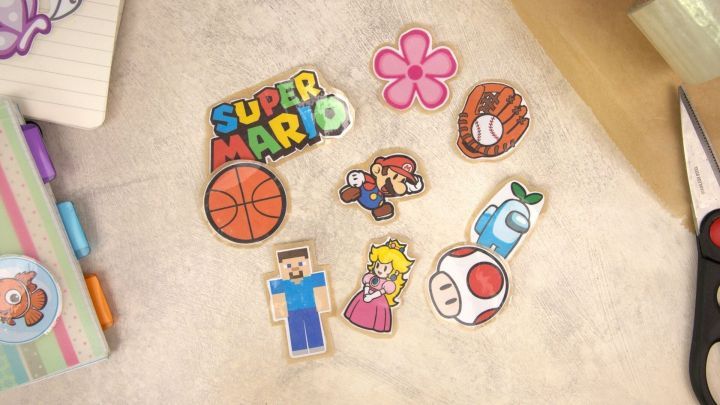 Bunte Sticker, darunter Super Mario, Basketball und eine Blume, liegen auf einem hellen Untergrund neben B&uuml;robedarf.