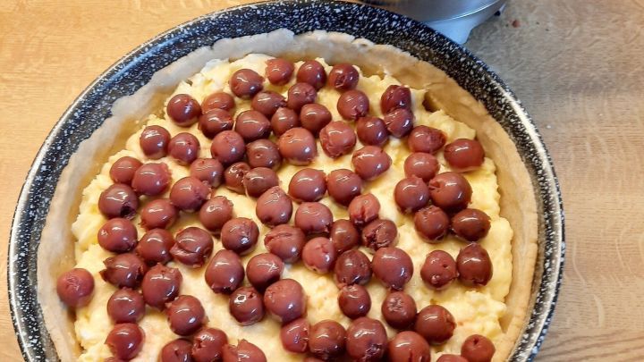 Der noch ungebackene Kuchen wird mit Kirschen belegt. Daneben steht eine Edelstahlsch&uuml;ssel mit Streusel bereit.