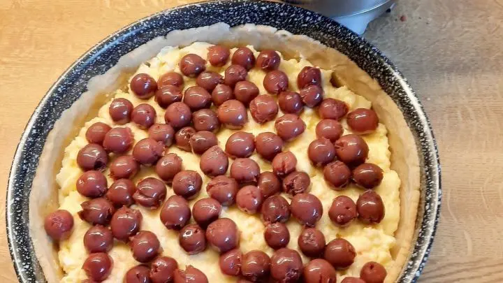 Der noch ungebackene Kuchen wird mit Kirschen belegt. Daneben steht eine Edelstahlsch&uuml;ssel mit Streusel bereit.
