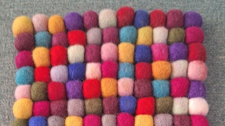 Ein bunter Filzuntersetzer liegt auf einem grauen Stoff, zeigt ein Muster aus kleinen Filzkugeln. Ger&uuml;che entfernen.