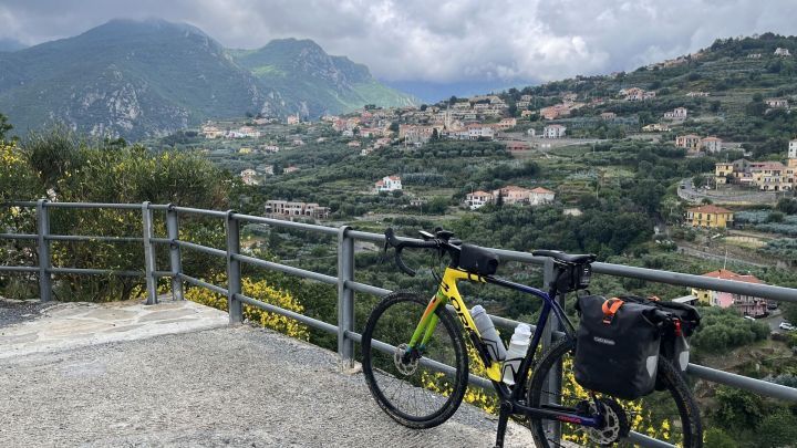 Ein Fahrrad steht auf einem Aussichtspunkt, mit Blick auf gr&uuml;ne H&uuml;gel und italienische Landschaft, w&auml;hrend Wolken vorbeiziehen.