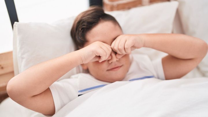 Ein Junge reibt sich die Augen, w&auml;hrend er im Bett liegt und frisch aufwacht, um den Schlafsand zu vertreiben.