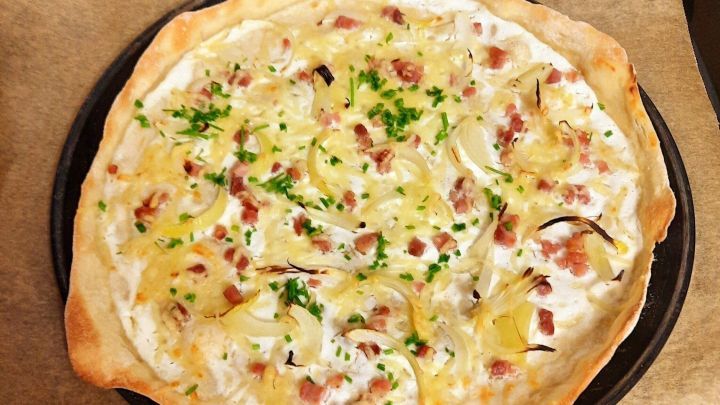 Ein frisch gebackener Flammkuchen liegt auf einem Backblech, belegt mit Zwiebeln, Schinken und K&auml;se.