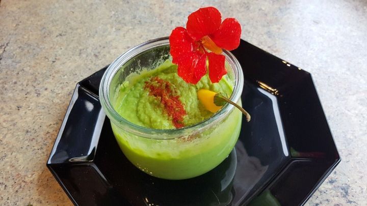 Leuchtend gr&uuml;ne Erbsen-Guacamole sitzt in einer Sch&uuml;ssel, dekoriert mit einer Bl&uuml;te und Paprika auf einem schwarzen Teller.