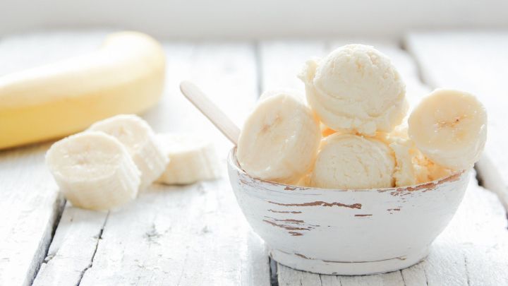 In einer wei&szlig;en Sch&uuml;ssel liegt cremiges Bananeneis, umgeben von frischen Bananenscheiben und einem ganzen Obstst&uuml;ck.