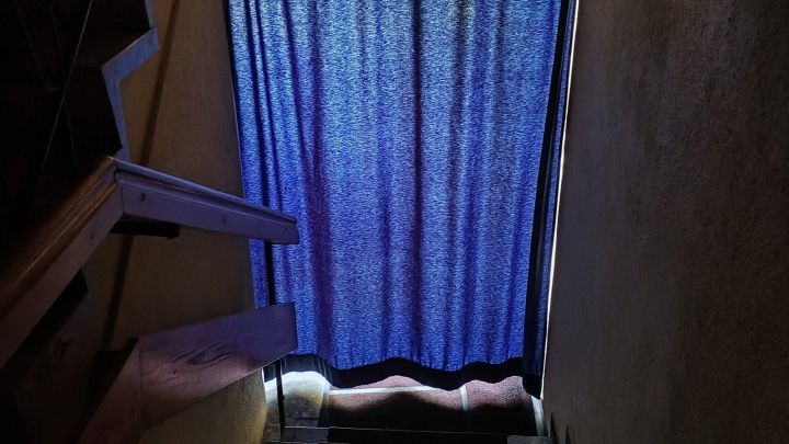 Ein blauer Vorhang h&auml;ngt an einer T&uuml;r, um die Treppe vor Licht und Hitze zu sch&uuml;tzen. Der Raum wirkt schattig und k&uuml;hl.