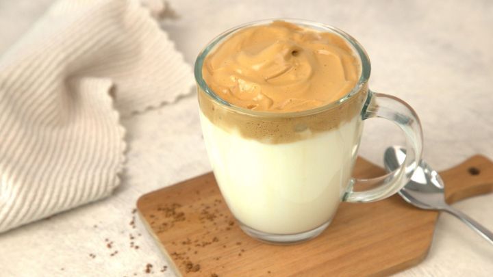 Ein Glas Dalgona Kaffee steht auf einem Holzbrett, oben schaumige Kaffeeschicht, daneben ein L&ouml;ffel.