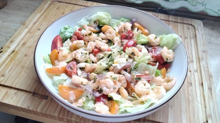 Eine bunte Sch&uuml;ssel mit Salat, Garnelen und Aprikosen wird appetitlich angerichtet auf einem Holzbrett pr&auml;sentiert.