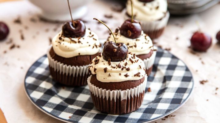 Schwarzw&auml;lder-Kirsch-Muffins stehen dekorativ auf einem karierten Teller, garniert mit Sahne, Kirschen und Schokoladensp&auml;nen.